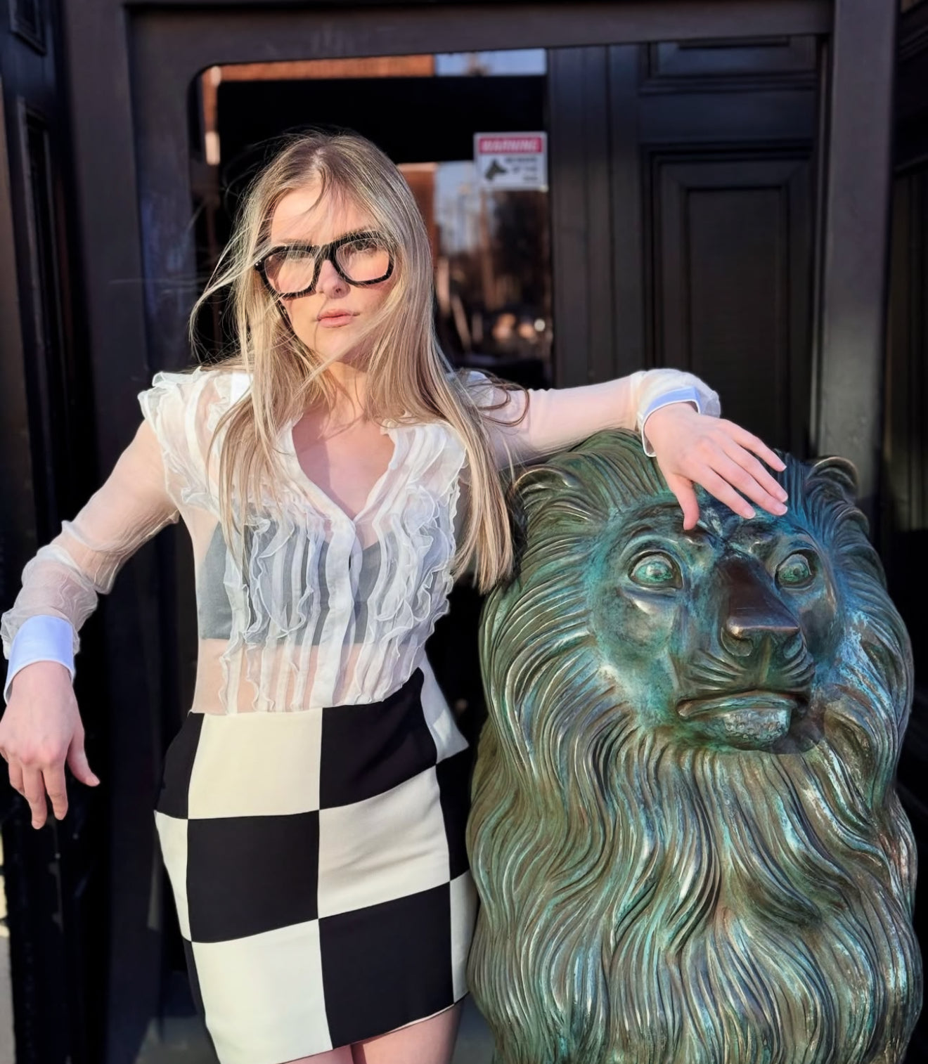 Woman posing with a lion statue in front of a building entrance while wearing a designer combo dress with sheer white top and black and white checkered skirt
