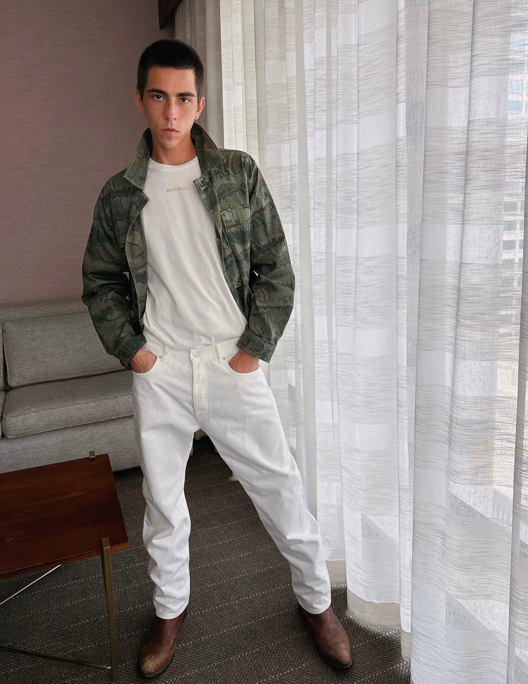 Male model wearing white designer pants and a green camo jacket standing in front of a white curtain with light coming through