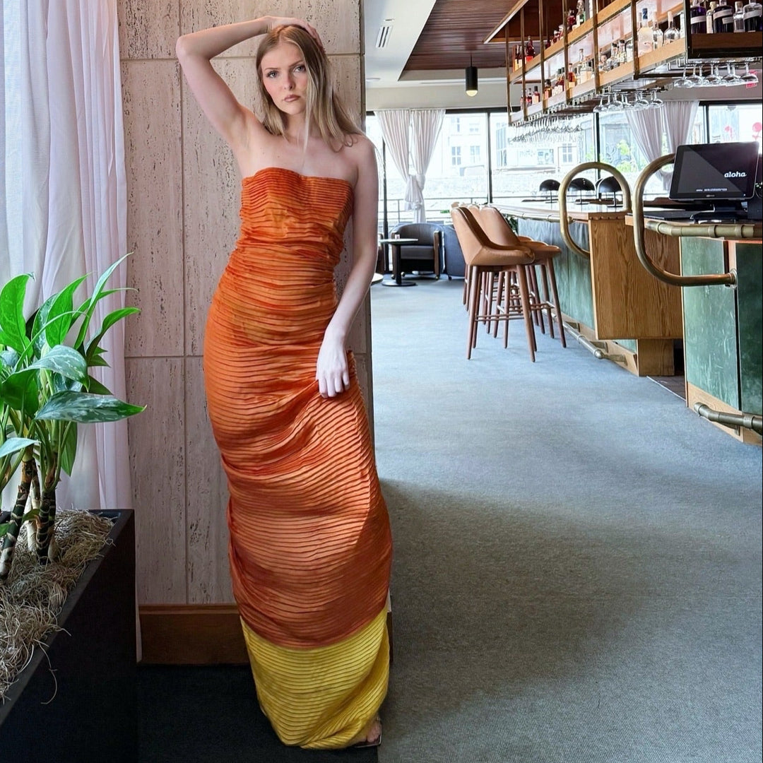 Woman in an orange designer dress standing in a modern interior setting with plants and a bar.