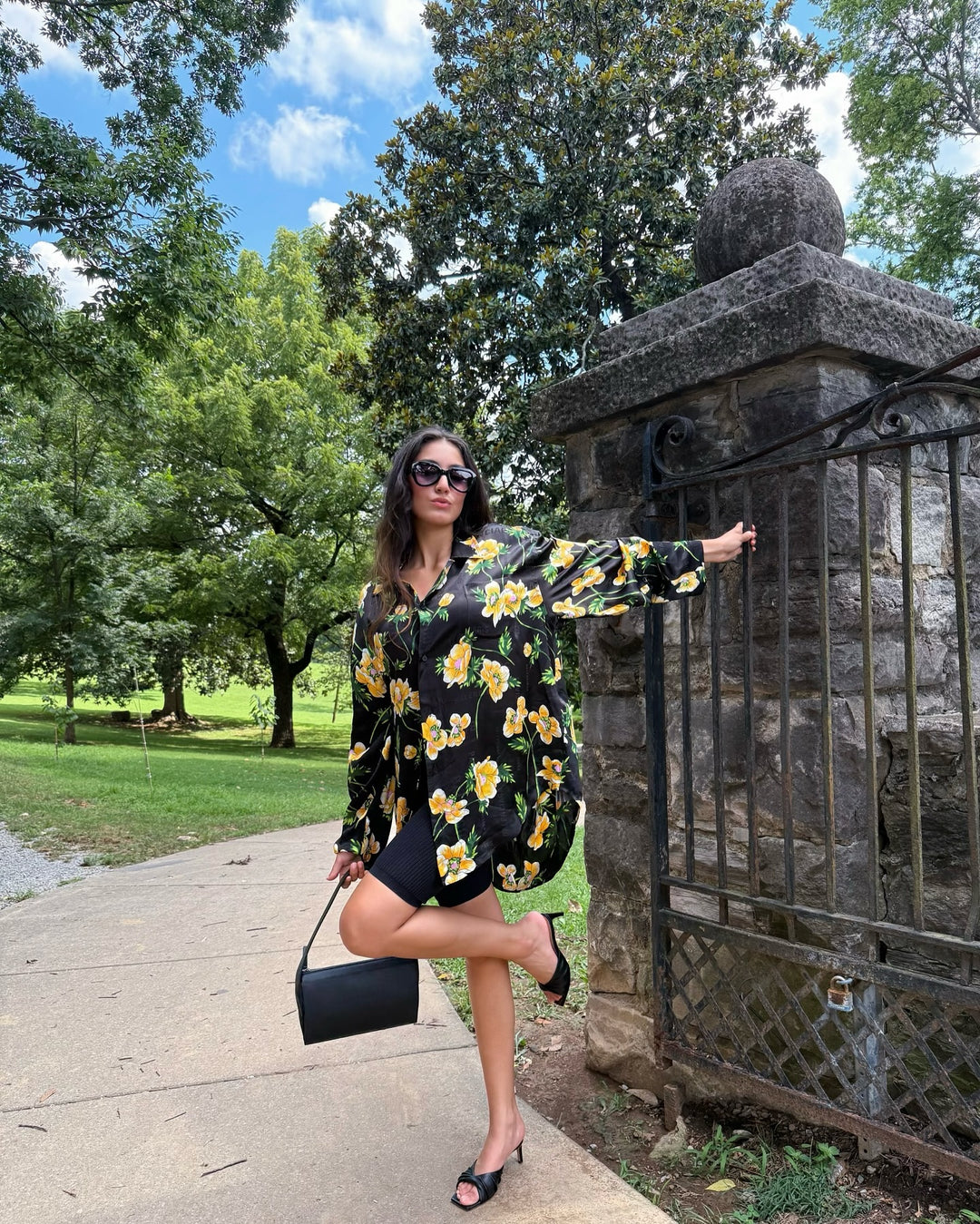 Female model in a park setting wearing Balenciaga black blouse with yellow flower print, black sunglasses, and a black designer handbag