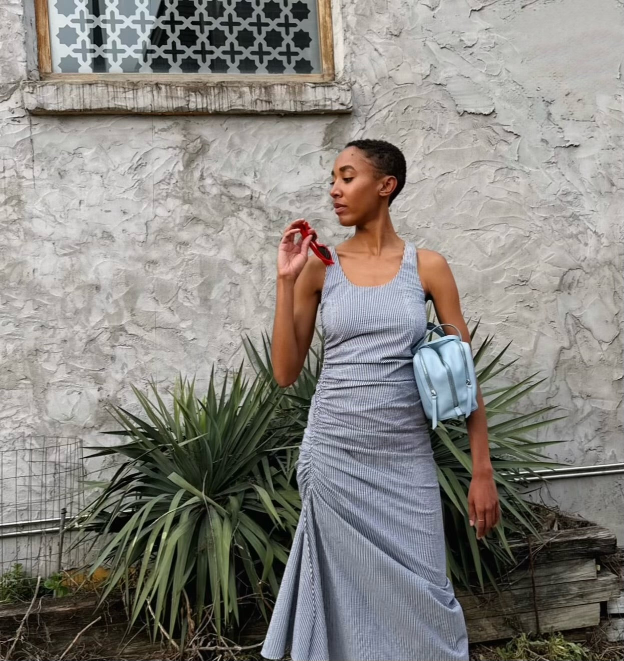 female model in designer black and white striped dress with a light blue designer handbag in front of a stone wall with green plants