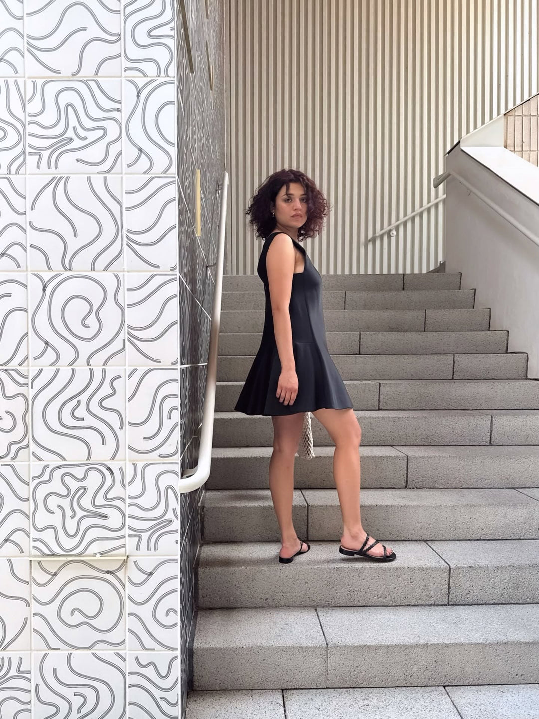female model wearing a black designer dress posing on a stone staircase with pattered tilework on the wall
