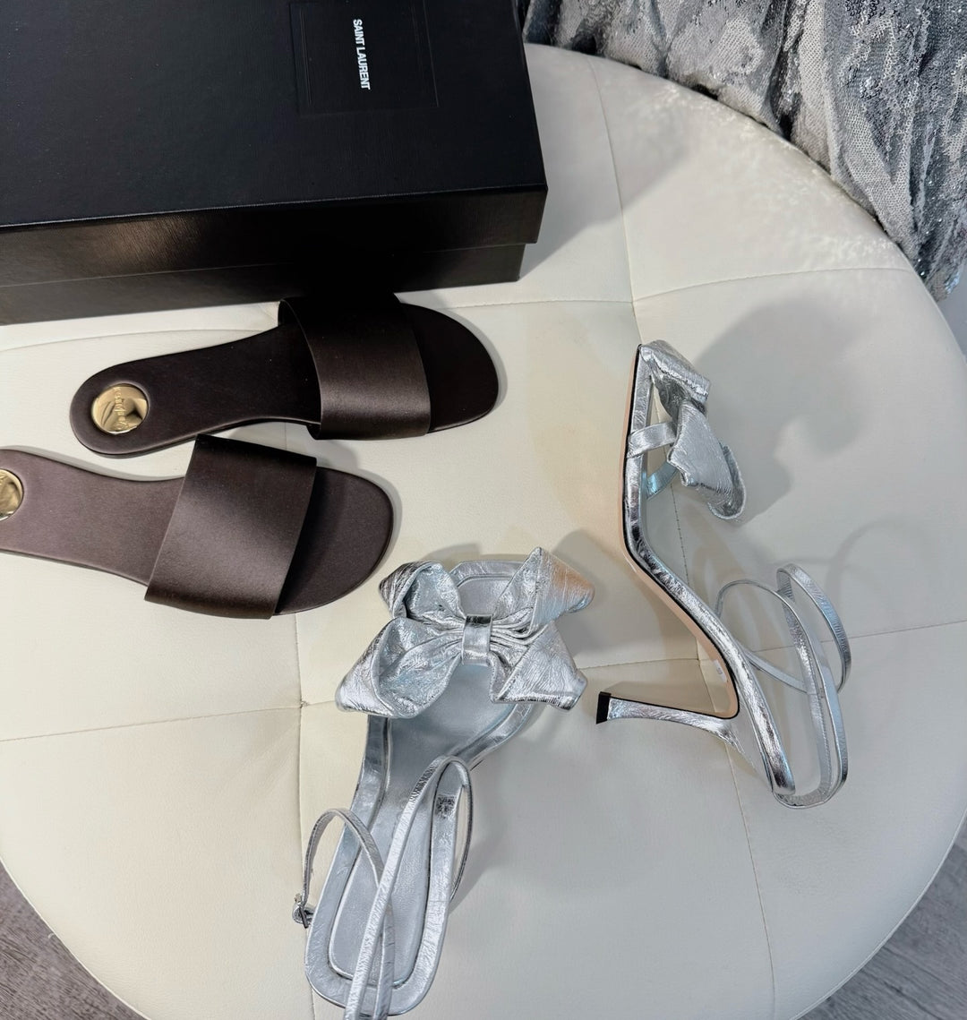 Brown designer sandal and silver high-heeled shoes on a white surface with a black Saint Laurent shoe box.