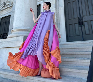 Female model standing in front of marble building while wearing flowy oversized purple and pink dress with orange ruffles