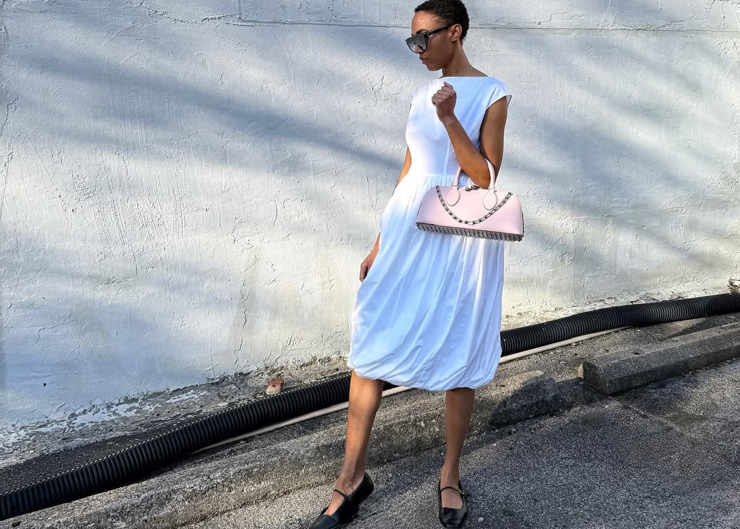 female model against rugged white cement wall and wearing a white designer dress with black sunglasses and a pink Valentino bag