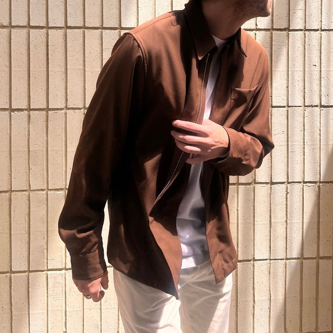 male model in front of yellow brick wall with focus on his brown designer zip front shirt with white t-shirt peeking through  and off-white jeans