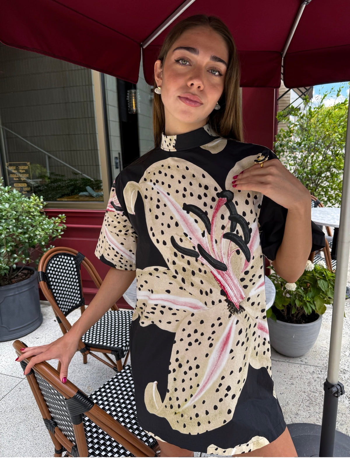 A woman wearing a short-sleeve black dress with a large beige floral print, black polka dots, and pink accents, posing on an outdoor patio under a red umbrella.