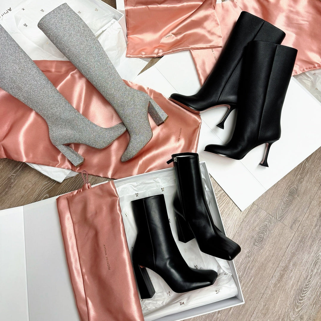 High-heeled boots in black leather and silver glitter on pink satin dust bags on a wooden floor.