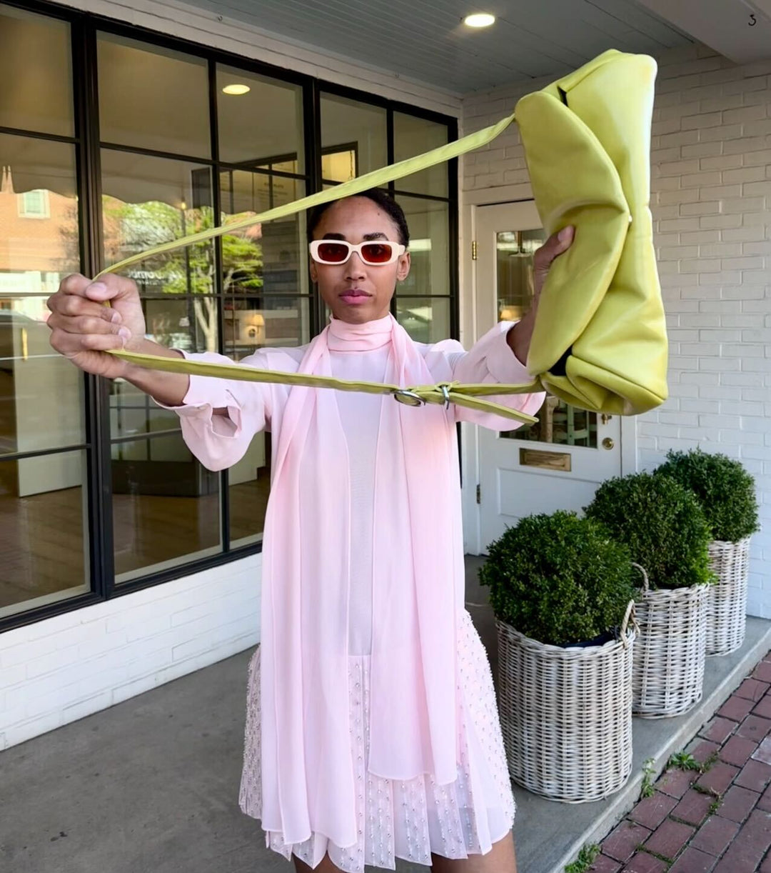 female model wearing a pretty pastel pink chiffon designer dress and pastel pink sunglasses while holding out a lime green bag in front of her towards the camera