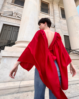 Female model facing away from camera wearing blue jeans and red draped top with deep v-neck at back