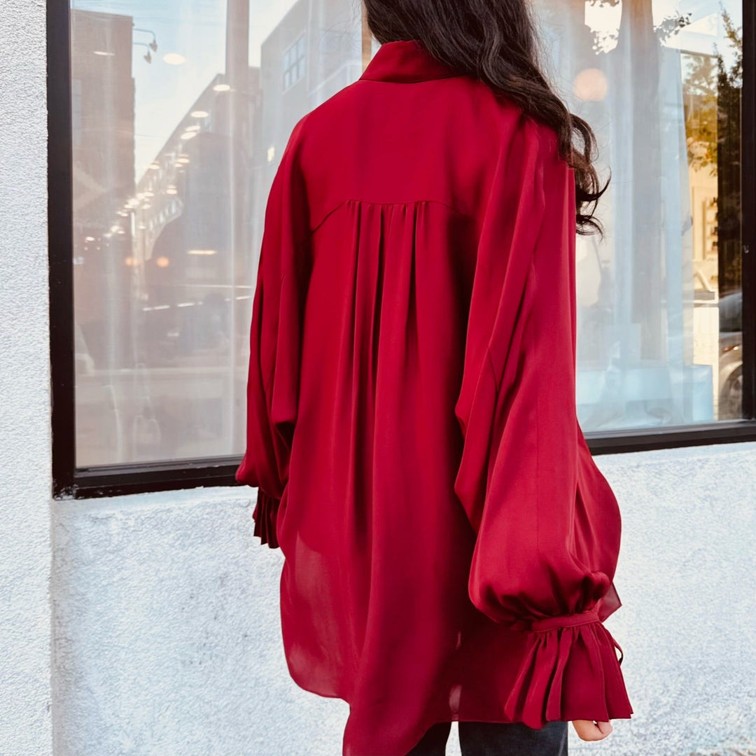 Female model wearing a red designer silk blouse with long sleeves in front of a window.