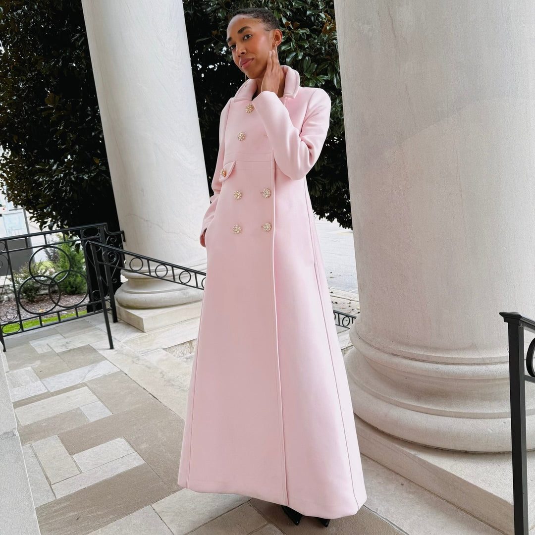 Female model wearing a pink designer fill-length wool coat standing between two large columns outdoors.