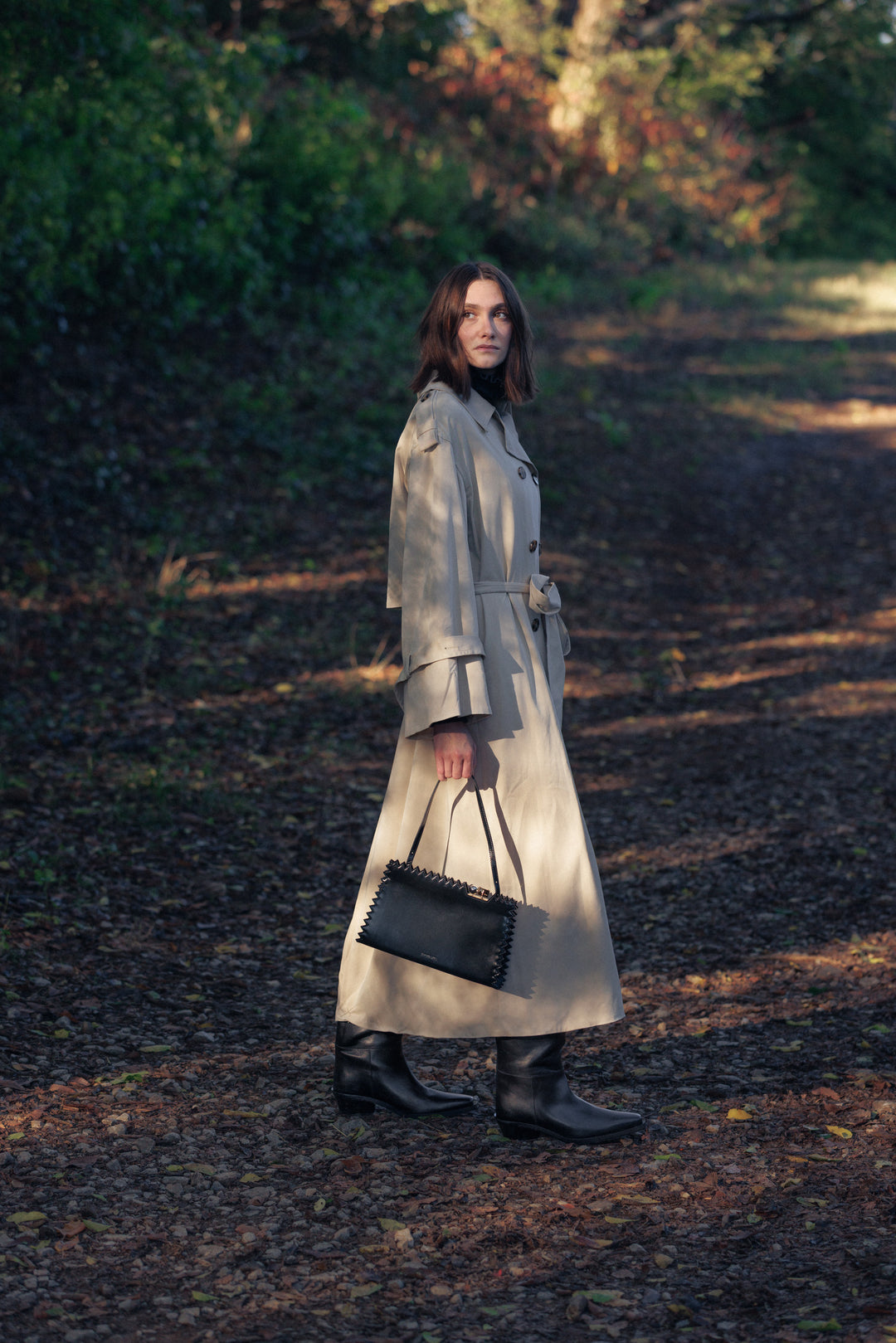Female model in a fall forest setting wearing a designer khaki trench coat and carrying a black designer handbag