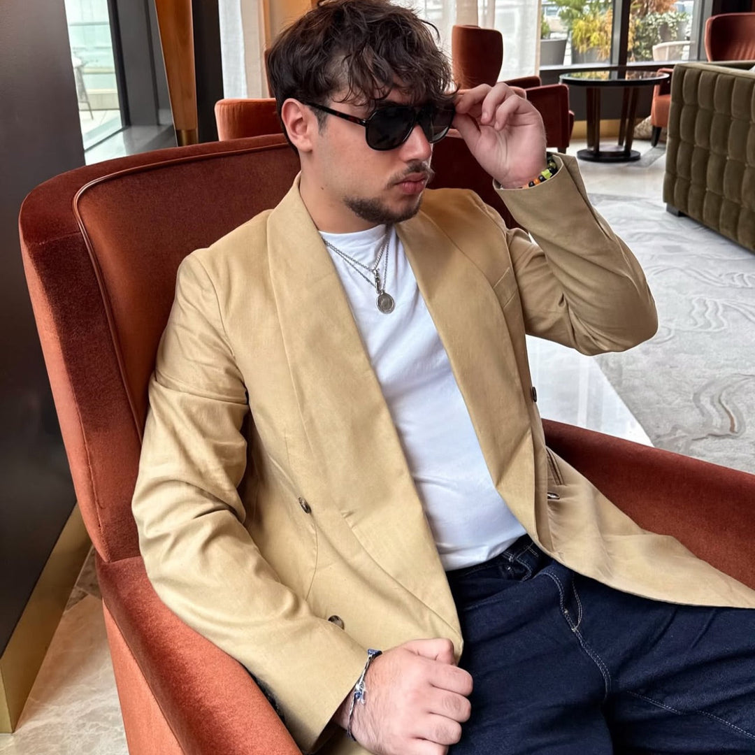 Man wearing a beige designer blazer and sunglasses sitting on a brown chair in a modern interior setting.