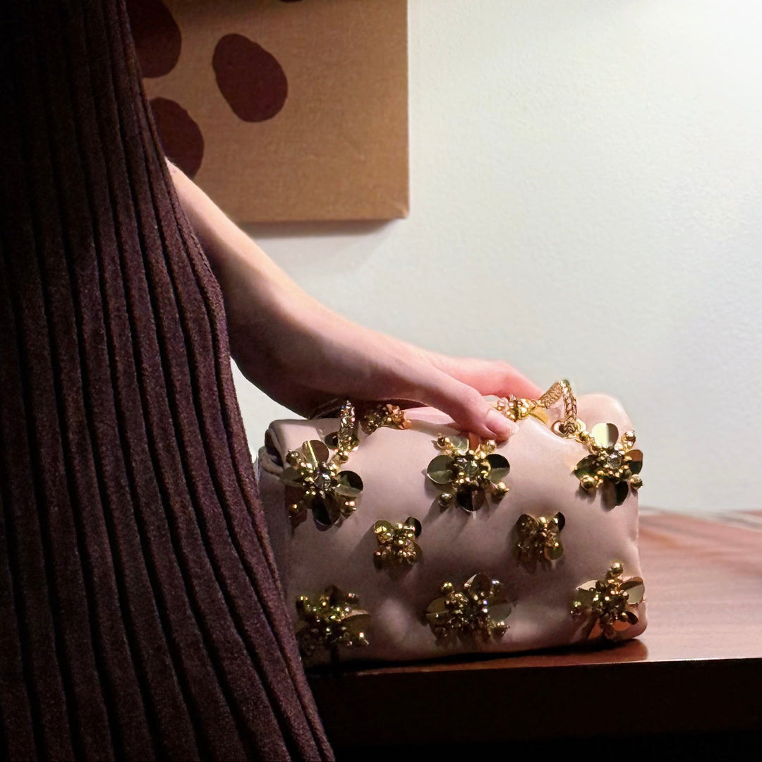 A person in a dark ribbed dress rests their hand on a blush pink clutch bag adorned with gold floral embellishments, set on a wooden table next to a brass lamp.