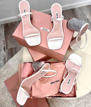 Top view of Miu Miu sandals in strappy white leather with chunky silver glitter heel and all laying across the pink Miu Miu boxes.