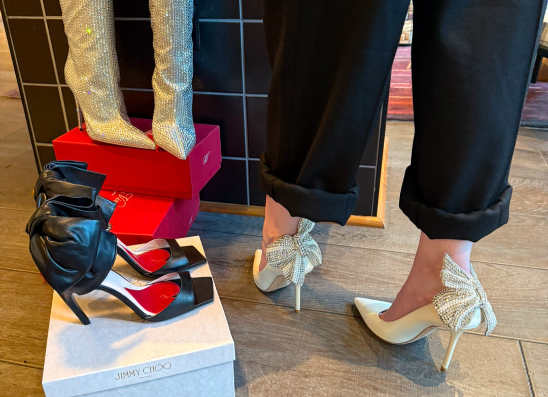 designer shoes on display including rhinestone-covered tall boots in cream, black heel sandals with a bow back, and our model wearing some white pumps with a crystal-covered bow back