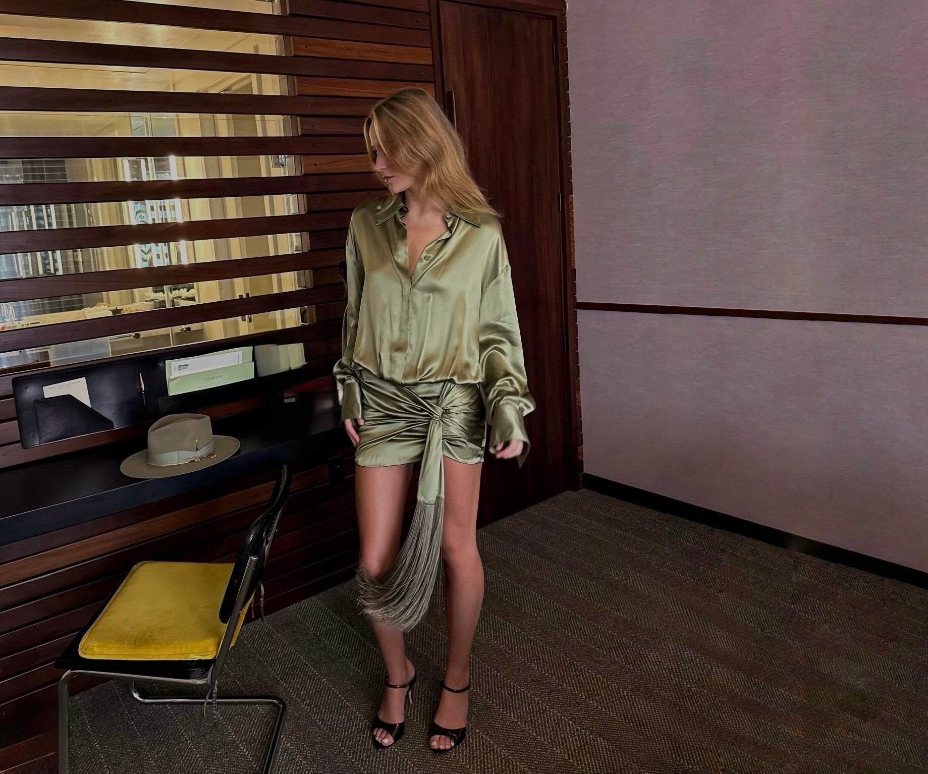A woman models a olive green satin mini dress with a draped tassel detail, standing in a modern room with wood paneling and a desk.
