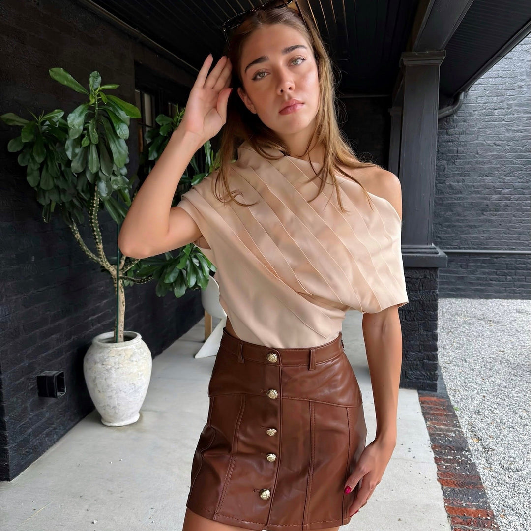 A woman posing on a covered walkway wearing a sheer, off-the-shoulder beige top and a brown faux-leather mini skirt with front buttons.